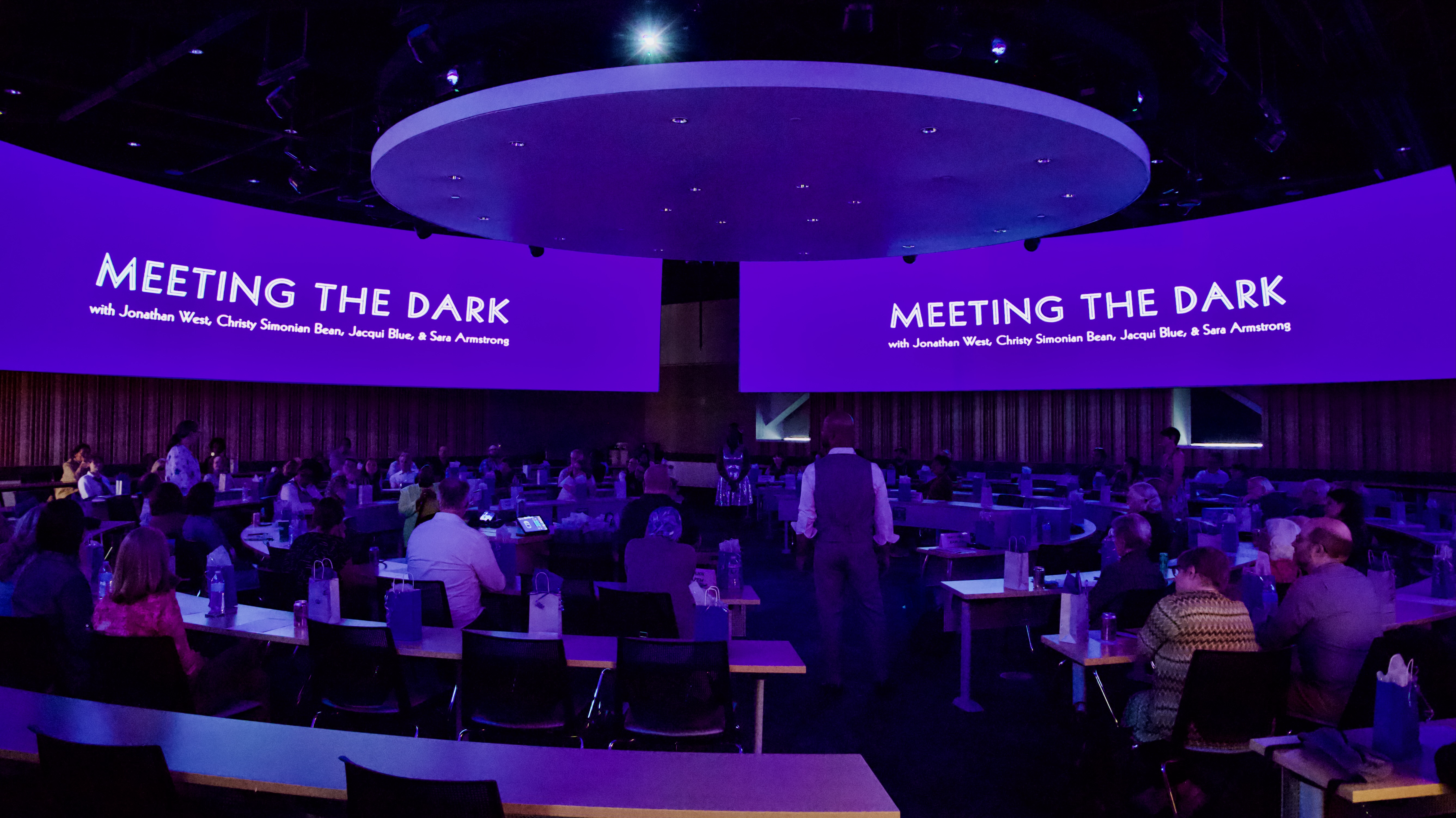 Audience in a circular auditorium and two screens that say "Meeting the Dark"