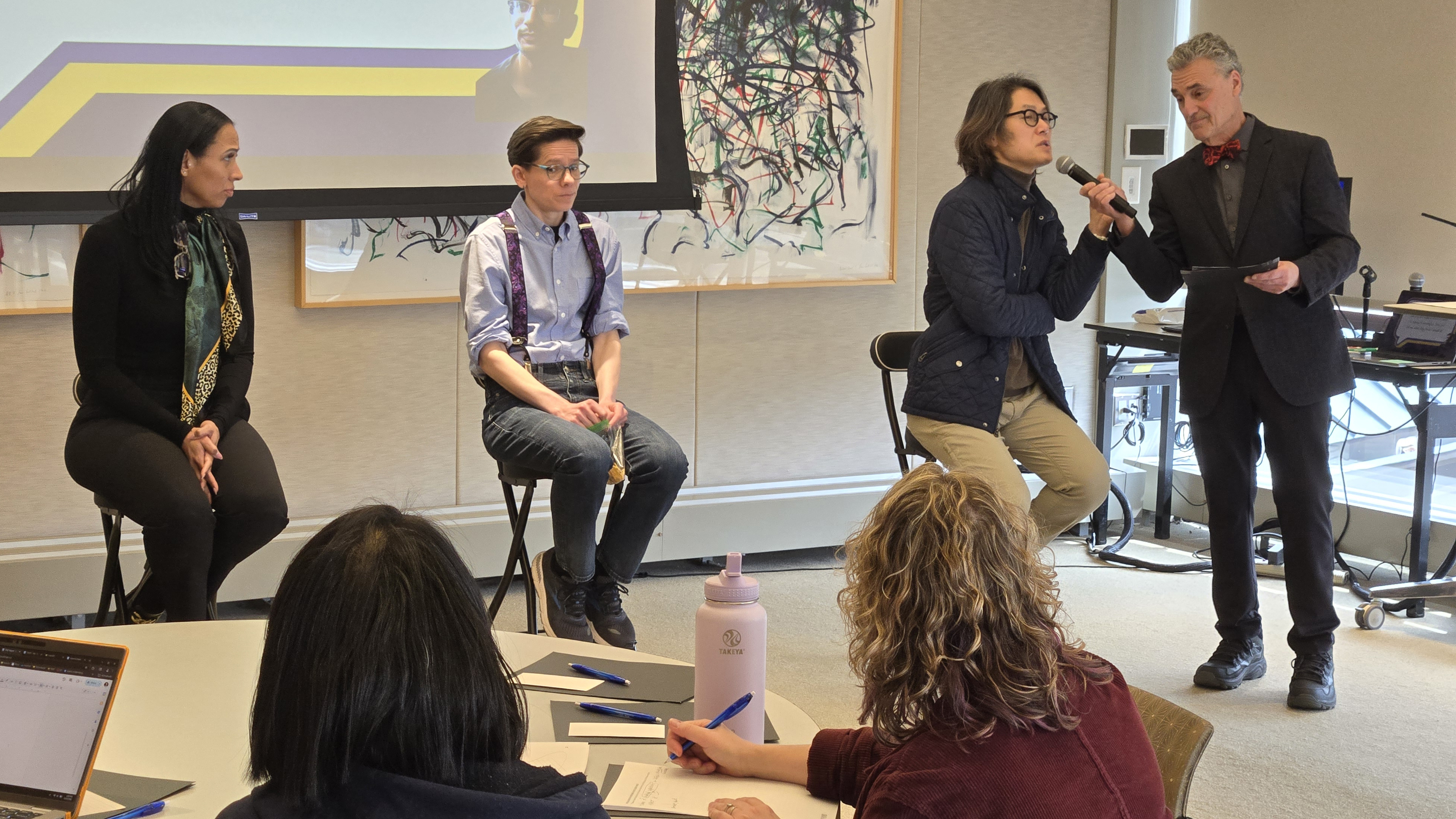 3 actors seating on stools while another stands and holds a microphone